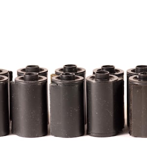 Puede incluir: Una fila de diez cartuchos de película negros, algunos con una etiqueta dorada, dispuestos en línea sobre un fondo blanco.