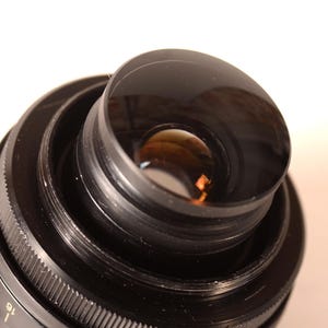 May include: Close-up of a black camera lens, featuring multiple concentric rings and a clear glass element. The lens has a ridged focus ring and a reflective interior, indicating a high-quality photographic device.