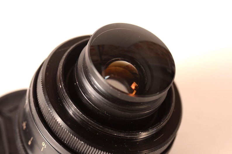May include: Close-up of a black camera lens, featuring multiple concentric rings and a clear glass element. The lens has a ridged focus ring and a reflective interior, indicating a high-quality photographic device.