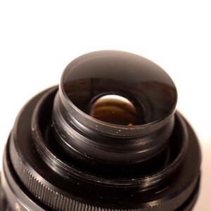 May include: Close-up of a black camera lens with a curved, reflective front element. The lens has concentric rings and a ridged focus ring. The background is plain white.