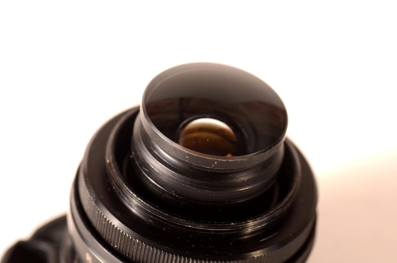 May include: Close-up of a black camera lens with a curved, reflective front element. The lens has concentric rings and a ridged focus ring. The background is plain white.