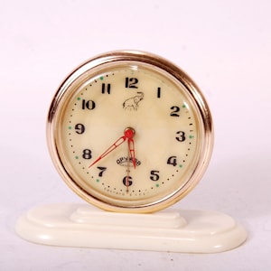 Può includere: Sveglia vintage rotonda con quadrante color crema e lunetta dorata. L'orologio presenta numeri neri, lancette rosse e un logo a forma di elefante. Poggia su una base color crema. Il diametro è di circa 7 cm.