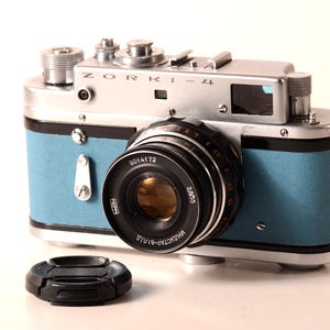 May include: A vintage Zorki-4 camera with a silver body and light blue leatherette accents. The camera has a black lens and a black lens cap. The text "ZORKI-4" is visible on the top of the camera.