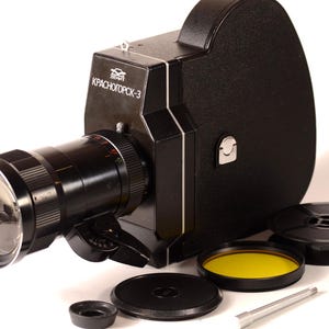 May include: A black Krasnogorsk-3 movie camera with a large lens, various accessories, and the text "KPACHOГOPCK-3". The camera is accompanied by a yellow filter, lens cap, and film reels, all set against a white background.