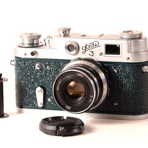 May include: Vintage silver and green camera with a lens, film canister, and lens cap. The camera has the Cyrillic text "Зоркий 3" on the top. The camera body is green with a textured finish. The lens is black and silver.