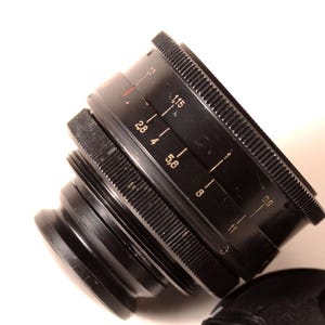 May include: Black camera lens with white markings, including f-stop numbers. The lens has a ridged focus ring and a lens cap. The lens is angled, showing the details of the aperture settings.