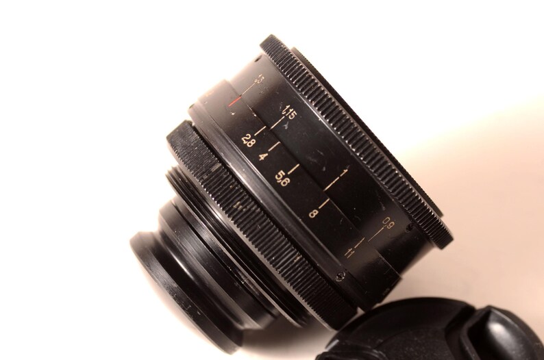 May include: Black camera lens with white markings, including f-stop numbers. The lens has a ridged focus ring and a lens cap. The lens is angled, showing the details of the aperture settings.