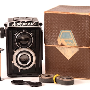 Puede incluir: Una cámara réflex de doble lente negra vintage con la palabra "Любитель" en la parte superior, acompañada de una caja marrón, un rollo de película, una tapa de lente y una correa de cuero. La cámara tiene dos lentes y un borde plateado.
