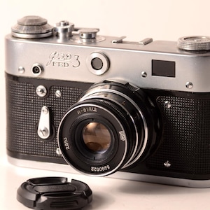 May include: A vintage FED 3 camera with a silver top and black textured body. The lens is black with silver accents, and a lens cap sits in front. The camera has the text "FED 3" on the front.