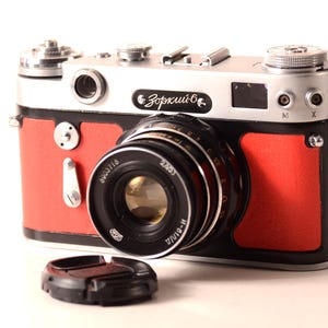 May include: A vintage camera with a red and silver body and a black lens. The camera has the word "Зоркий" printed on the front. A black lens cap sits in front of the camera.