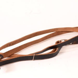 May include: A brown leather camera strap with a black leather strap attached to it. The strap has a metal buckle and a metal loop.