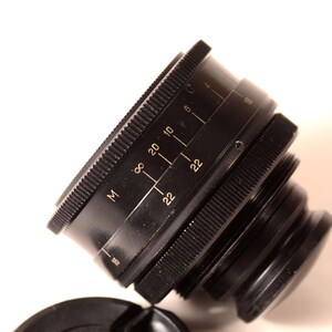 May include: A black camera lens with a ridged focus ring and aperture markings. The lens has a series of numbers and the letter "M" etched on its side. The lens cap is visible in the foreground.