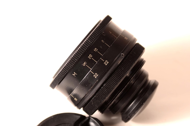 May include: A black camera lens with a ridged focus ring and aperture markings. The lens has a series of numbers and the letter "M" etched on its side. The lens cap is visible in the foreground.
