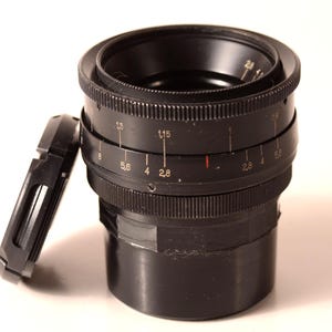 May include: A black camera lens with a detached lens cap. The lens features rings with numerical markings, including f-stops. The lens cap is rectangular and black. The background is white.