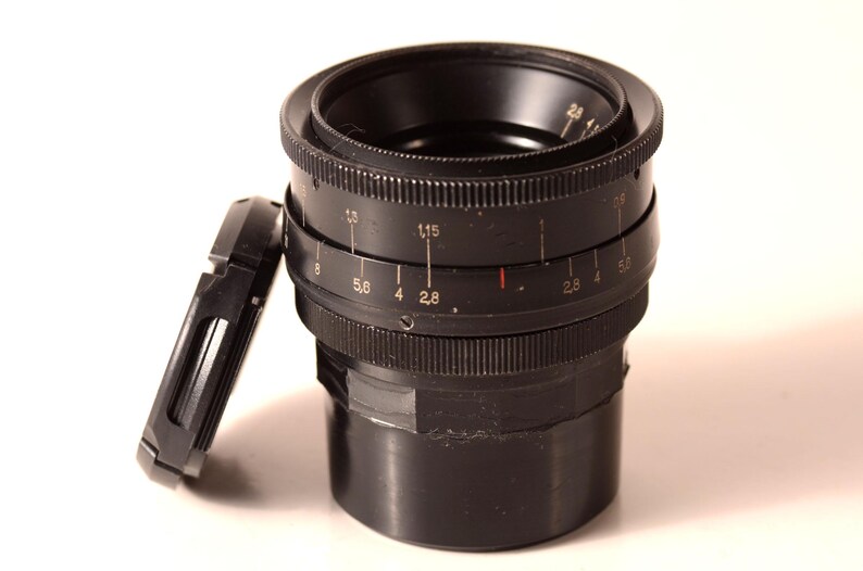 May include: A black camera lens with a detached lens cap. The lens features rings with numerical markings, including f-stops. The lens cap is rectangular and black. The background is white.