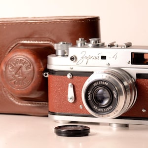 Può includere: Una fotocamera a telemetro Zorki-4 con corpo color argento e marrone, dotata di un obiettivo con etichetta Industar-50 e testo in cirillico. La fotocamera è accompagnata da una custodia in pelle marrone con logo in rilievo e un tappo nero.