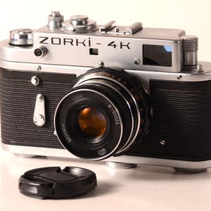Op de afbeelding: Een vintage Zorki-4K camera met een zilveren behuizing en zwarte getextureerde grip. De lens is zwart met een goudkleurig interieur. Een zwarte lensdop ligt voor de camera. De tekst "ZORKI-4K" is zichtbaar.