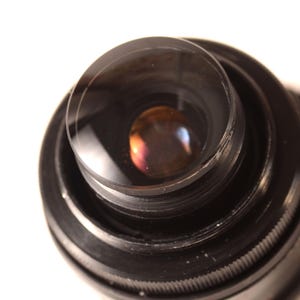 May include: A close-up shot of a black camera lens, featuring multiple concentric rings. The lens has a clear, convex glass element that reflects a spectrum of colours. The background is plain white.