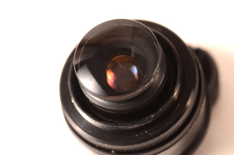 May include: A close-up shot of a black camera lens, featuring multiple concentric rings. The lens has a clear, convex glass element that reflects a spectrum of colours. The background is plain white.