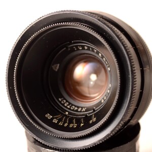May include: A close-up shot of a black Jupiter-8 2/50 camera lens. The lens features various markings and numbers, including "Jupiter-8" and "2/50". The lens is set against a white backdrop.