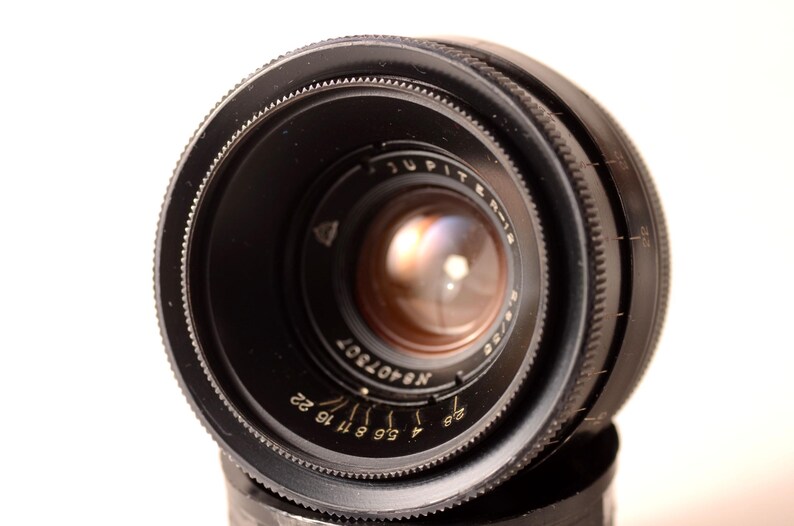 May include: A close-up shot of a black Jupiter-8 2/50 camera lens. The lens features various markings and numbers, including "Jupiter-8" and "2/50". The lens is set against a white backdrop.