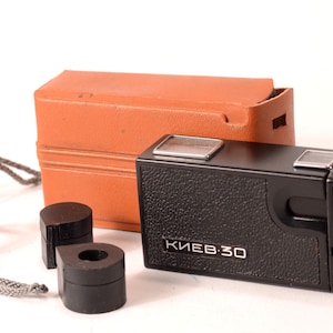 May include: A black Kiev-30 camera with a brown leather case and two black lens caps. The camera has a silver inscription that reads "Kiev-30".