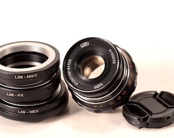 May include: A black camera lens with a silver ring and a black lens cap. The lens has the text "INDUSTAR-61Л/Д 2,8/55" and "9160033" printed on it. The lens is surrounded by a stack of black camera lens adapters with the text "L39 - M4/3", "L39 - FX", and "L39 - NEX" printed on them.