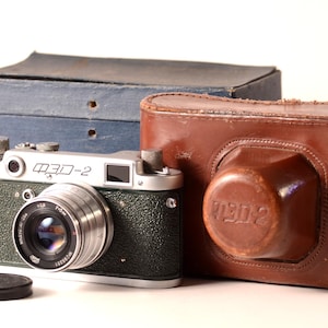 Puede incluir: Cámara FED-2 vintage con funda de cuero y tapa de objetivo. El cuerpo de la cámara es verde y plateado, con el nombre del modelo impreso en la parte superior. La funda de cuero marrón está grabada con el nombre del modelo. Una caja azul está detrás de la cámara.