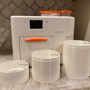 May include: A white Rotimatic appliance with an orange handle and the word "rotimatic" printed on the front. Three white plastic containers are in front of the appliance. The containers measure 2.4 inches, 4.4 inches, and 6.4 inches in height.