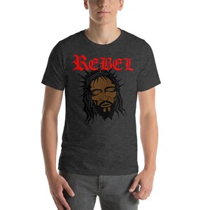 May include: A dark heather grey t-shirt with a black and brown image of a person with a crown of thorns and the word "REBEL" in red lettering.