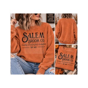 May include: A burnt orange long-sleeve crewneck sweatshirt featuring black text that reads Salem Broom Co. Quality Handcrafted Enchanted EST 1692. The garment has a distressed, vintage-style wash and a ribbed collar, cuffs, and hemline.