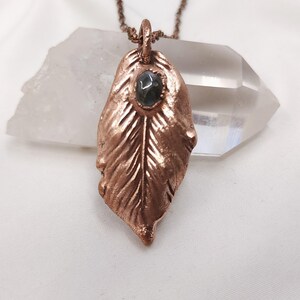 May include: A copper-colored pendant necklace featuring a leaf design with intricate detailing. The leaf pendant has a small, oval-shaped, dark green stone in the center. The necklace chain is a matching copper color. The jewelry is displayed on a white surface.