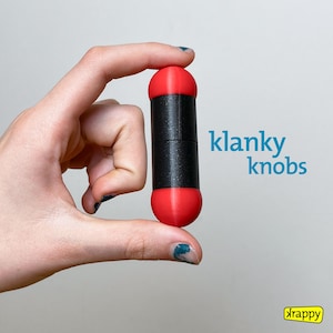 Peut inclure: Une main tient un objet cylindrique rouge et noir avec le texte "klanky knobs" imprimé sur un fond blanc. L'objet est en plastique et a une surface texturée.