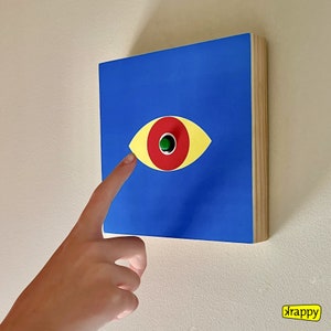 May include: A blue square with a yellow, red, and green eye design. A finger is pointing at the eye.