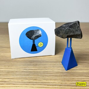May include: A blue plastic rock holder with a grey rock balanced on top. The holder has a triangular base and two prongs that hold the rock in place. The text "rock holder" is printed on a yellow circle on the box.