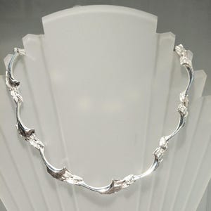 May include: A silver-toned necklace with an organic, flowing design. The necklace features irregular, textured elements along its curved structure, creating a unique and artistic aesthetic. The necklace is displayed on a white mannequin.