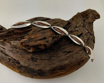 May include: A silver bracelet featuring a series of elongated, oval-shaped links. Each link has a polished, reflective surface with a dark, reddish-brown interior. The bracelet is displayed on a piece of weathered, dark brown wood.