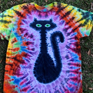May include: A tie-dye t-shirt featuring a black cat design with green eyes. The shirt has a spiral pattern in shades of orange, yellow, purple, and blue. The cat's silhouette is centered on the front of the shirt.