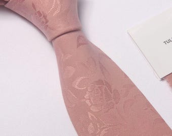 May include: A dusty rose necktie featuring a tonal jacquard floral pattern with visible leaves and blossoms. The tie is knotted at the top and includes a partial white tag with the visible text TUL. The fabric has a subtle sheen and texture.