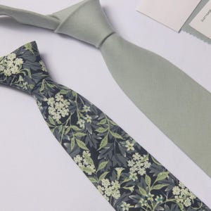 May include: Two neckties displayed together. One tie is a solid, muted sage green colour with a textured fabric finish. The second tie features a dark charcoal background with a dense, repeating pattern of light green leaves and small white flowers.