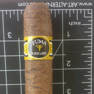 May include: A brown cigar with a gold band that says "FUMA BLACK LABEL". The cigar is about 4 inches long and is sitting on a black grid.