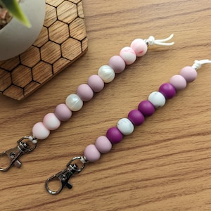 Peut inclure: Deux porte-clés perlés avec des fermoirs argentés. Un porte-clés est rose et blanc avec une perle. L'autre porte-clés est violet et blanc avec une perle marbrée.