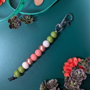 Puede incluir: Un llavero de cuentas con un cierre plateado. Las cuentas son en tonos de verde, blanco, rosa y cobre. El llavero se muestra sobre una superficie verde oscuro, con flores decorativas y un dije de madera en forma de bellota.