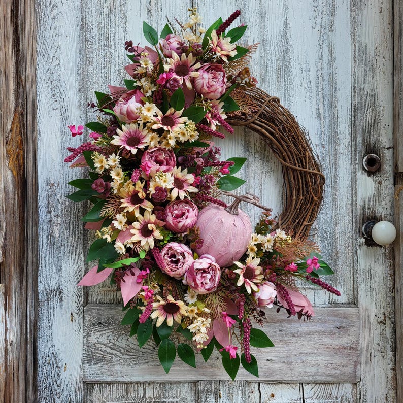 May include: A pink and green floral wreath with a pink pumpkin in the center. The wreath is made of artificial flowers and greenery and is displayed on a white wooden door.