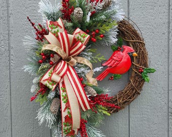 May include: A decorative grapevine wreath featuring a red cardinal, pinecones, and faux greenery. The wreath is adorned with a large bow with red and white stripes and a tan ribbon with cardinal designs. Red berries add a festive touch.