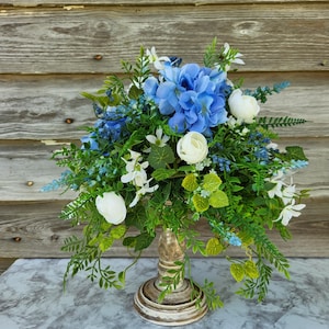 May include: A blue and white floral arrangement in a rustic wooden pedestal. The arrangement features blue hydrangeas, white flowers, and green foliage.