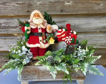 May include: A rustic wooden box centerpiece featuring a Santa Claus figurine holding a bag of gifts, a metal sleigh filled with presents, and a variety of greenery, including ferns and pine.
