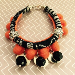 Puede incluir: Una pulsera artesanal con una banda estampada en blanco y negro, detalles en naranja y pompones. Incluye cuentas y dijes de metal plateado, además de cuentas facetadas negras. Un accesorio único y llamativo.