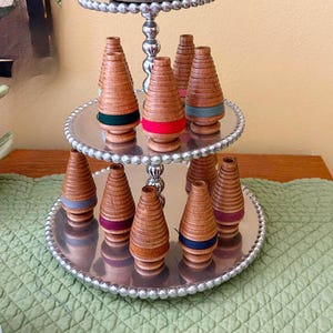 May include: A tiered silver serving tray displays several wooden spools with colorful bands. The spools are arranged on two levels, with a variety of colors including green, red, blue, and purple. The tray is set on a green quilted surface.