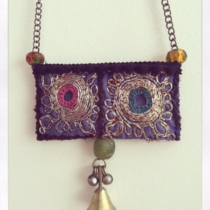 Puede incluir: Un collar hecho a mano con un colgante rectangular morado. El colgante presenta círculos bordados en rosa y azul, con detalles de hilo plateado. Una pequeña campana de latón y una cuenta verde cuelgan de la parte inferior. El collar tiene una cadena con cuentas decorativas.